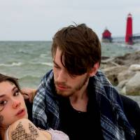 Photograph featuring two a male-presenting figure holding a female-presenting one on their arms at a lake shore